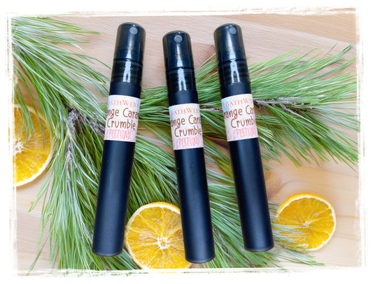 Three black Orange Caramel Crumble perfume spray bottles arranged with orange slices and pine branches.