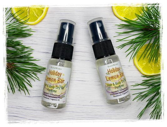 Two Holiday Lemon Bar Hair & Body Dry Oil spray bottles with clear caps, arranged with lemon slices and pine branches on white wood.