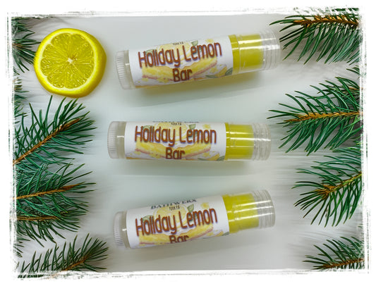 Three tubes of Holiday Lemon Bar lip balm arranged with a lemon slice and evergreen branches.