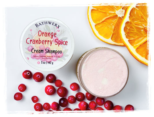 Orange Cranberry Spice cream shampoo in an open jar with pale pink cream, surrounded by cranberries and orange slices.