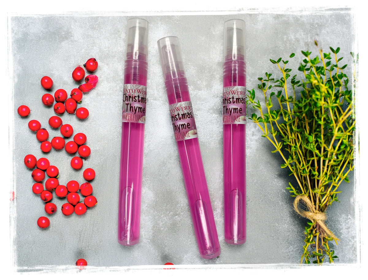 Three pink Christmas Thyme spray perfume bottles beside fresh thyme sprigs and red berries.