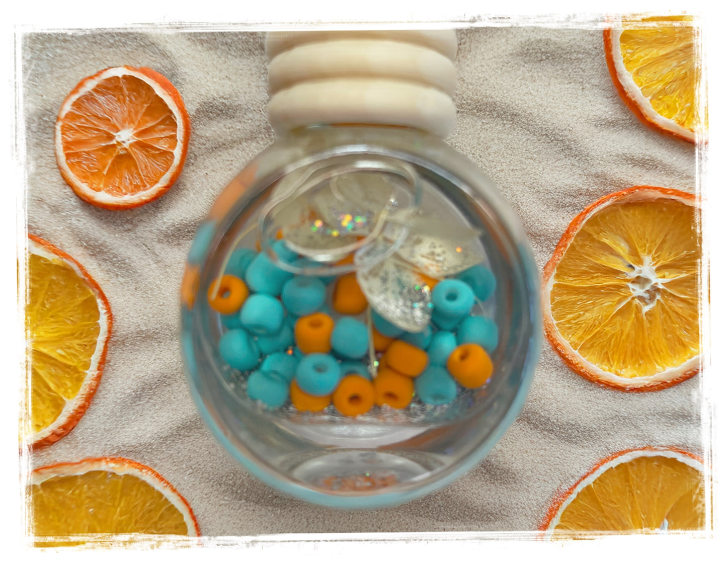 Round glass car charm with blue and orange beads on a sandy surface with orange slices around it.