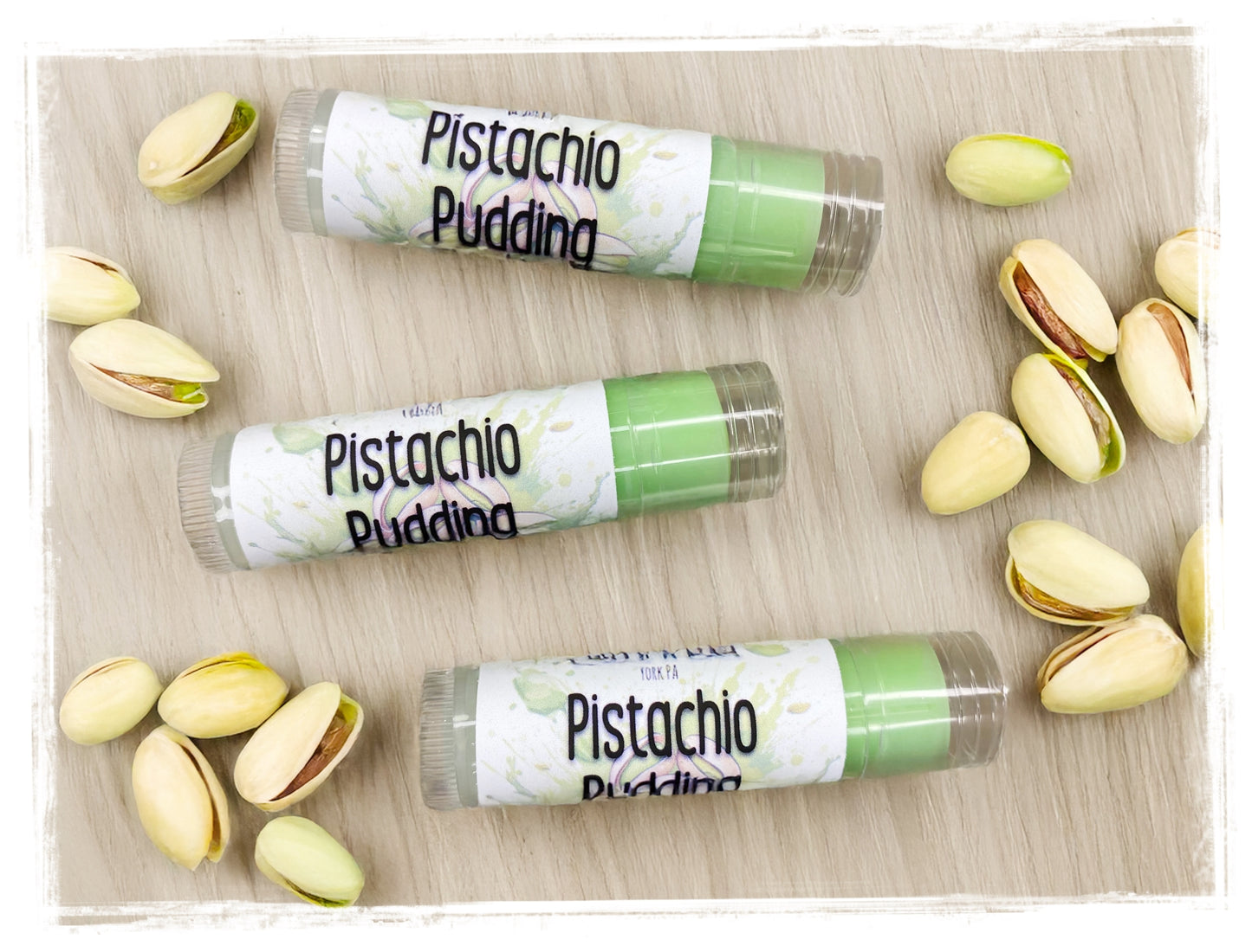 Three lip balm tubes labeled 'Pistachio Pudding' surrounded by pistachios on a wooden surface.