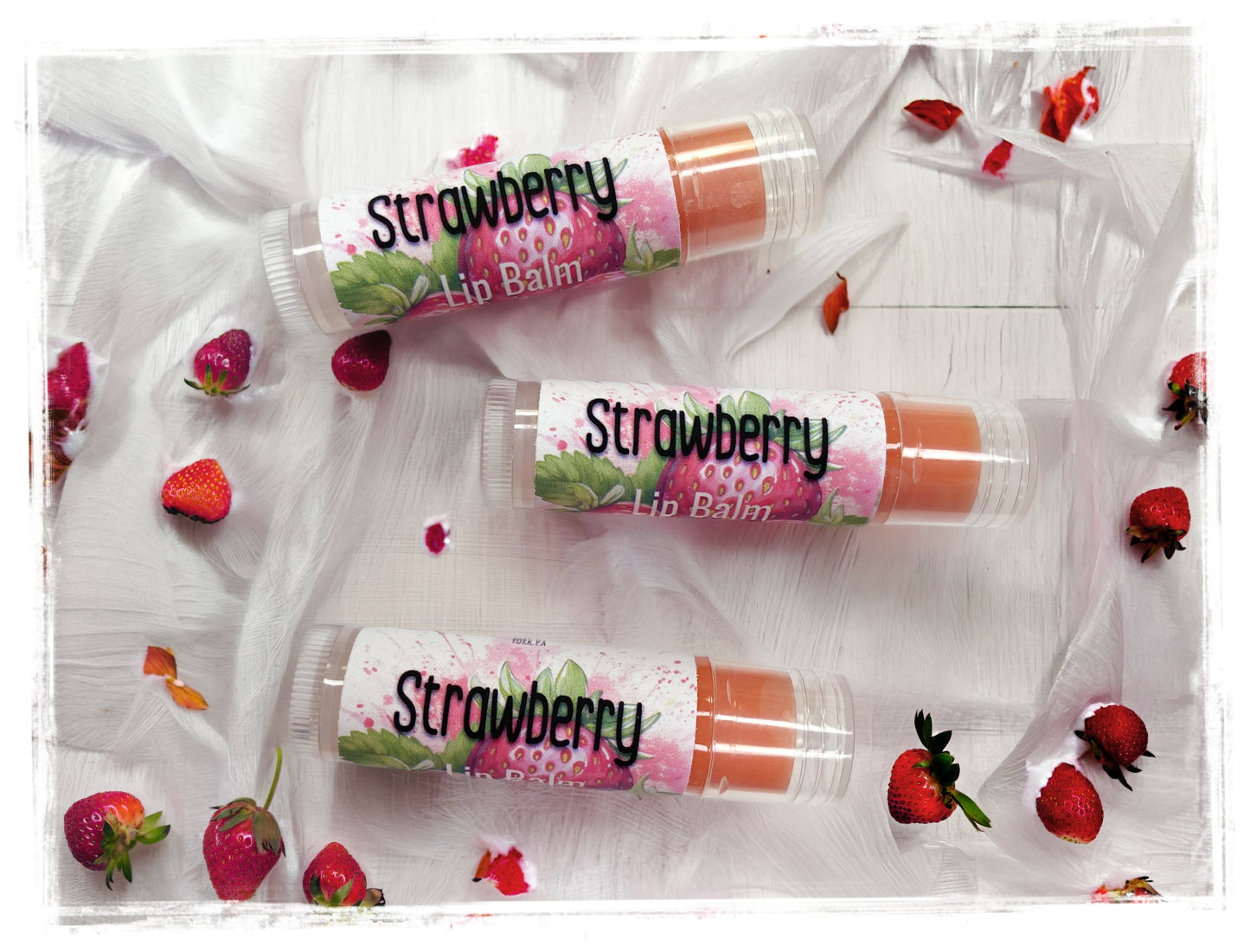 BathWerx Strawberry Lip Balm, three clear tubes with strawberry illustrated labels, surrounded by fresh strawberries and rose petals on white surface