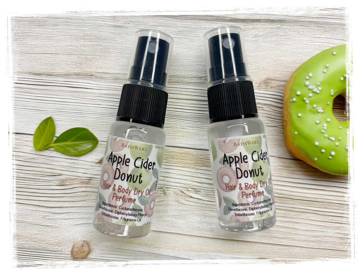 Two BathWerx Apple Cider Donut Hair & Body Dry Oil Perfume spray bottles on a white wood surface, styled with fresh green leaves and a green apple cider donut