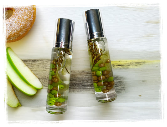 Two BathWerx Apple Cider Donut Roll-on Perfume bottles with silver caps, filled with oil, green apple beads, and botanicals, styled on a white wood surface with fresh green apple slices and a cinnamon donut
