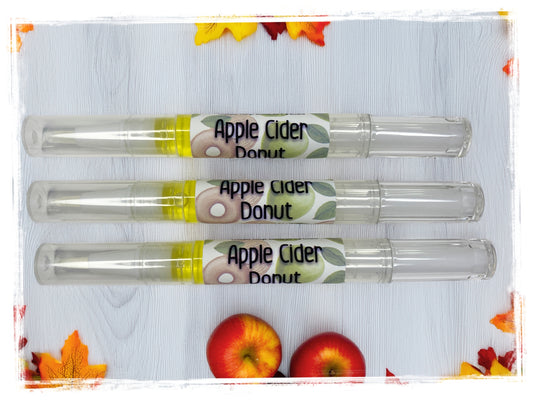 Three Apple Cider Donut Moisturizing Nail Pens displayed horizontally on a white wood surface, surrounded by autumn leaves and two red apples, each pen featuring a label with green apple and donut graphics