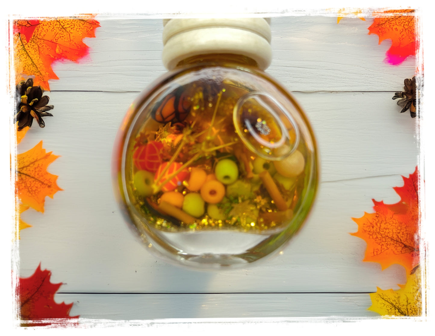 BathWerx Autumn Leaves scented car charm — a clear round ornament filled with fall botanicals, mini pumpkins, cinnamon sticks, green beads, and gold glitter, framed by colorful autumn leaves and pinecones on a rustic white wood surface.