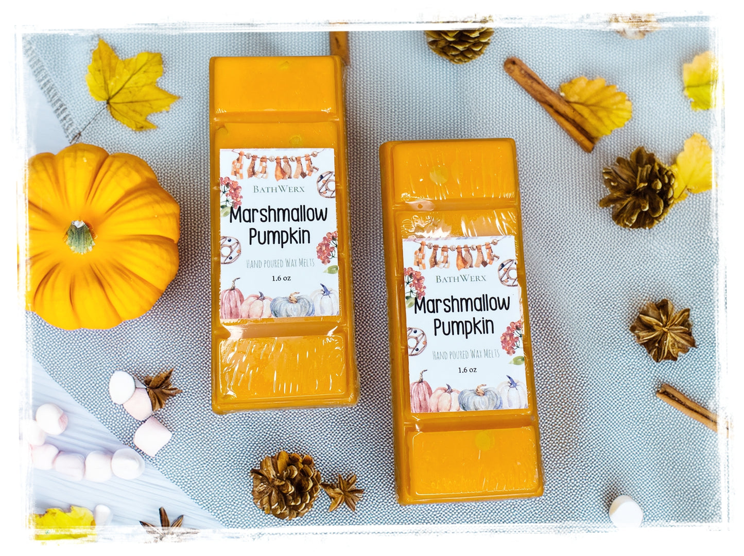Two BathWerx Marshmallow Pumpkin hand-poured wax melts in golden orange, displayed on a blue linen surface surrounded by autumn leaves, pine cones, star anise, cinnamon sticks, a mini pumpkin, and white marshmallows.