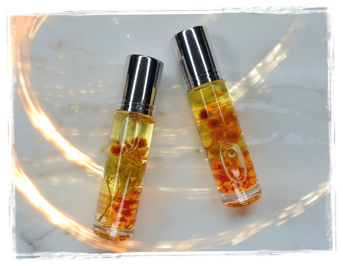 BathWerx Pumpkin Patchouli Fall Roll-On Perfume Oil clear glass vials with silver caps filled with golden oil and dried orange florals, on white marble with warm light reflections.
