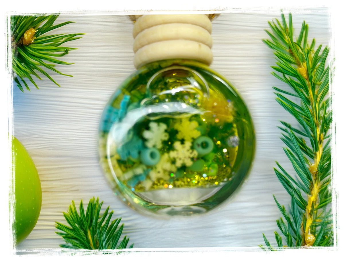 BathWerx Juniper + Apple Car Charm — clear glass ornament filled with green oil, white snowflake charms, teal ring beads, and gold glitter, displayed on a white wood surface with fresh pine branches and a green apple accent.