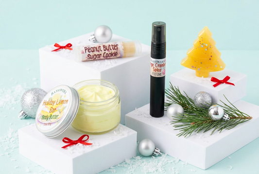 Holiday body butter, perfume, lip balm, and tree-shaped soap on white displays with silver ornaments and red bows against an icy aqua background.