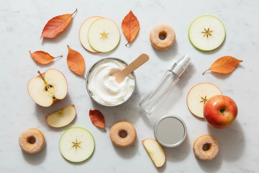 BathWerx body butter tin open to reveal fluffy whipped texture with a wooden spatula, paired with a clear plastic perfume spray bottle, arranged on a light marble surface with halved red apples, mini cinnamon-dusted donuts, and scattered fall leaves.