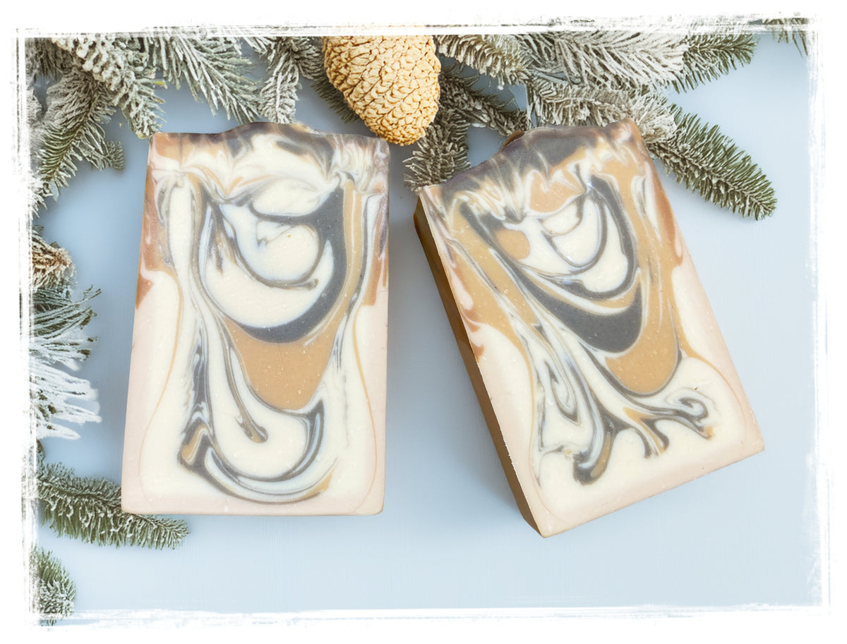 Two Cocoa Butter Cashmere goat milk soap bars with cream, tan, and brown swirls, framed by evergreen branches and a pinecone.