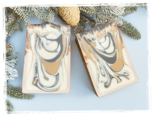 Two Cocoa Butter Cashmere goat milk soap bars with cream, tan, and brown swirls, framed by evergreen branches and a pinecone.