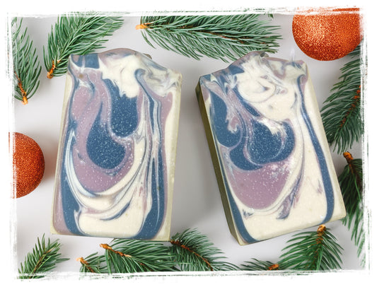Two Dark Woods goat milk soap bars with black, lavender, and cream swirls, styled with evergreen sprigs and orange ornaments.
