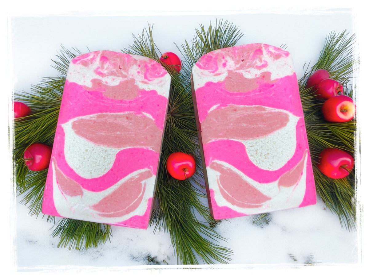 Two Winter Candy Apple goat milk soap bars with bright pink, blush, and white swirls, arranged on pine branches with small red apples.