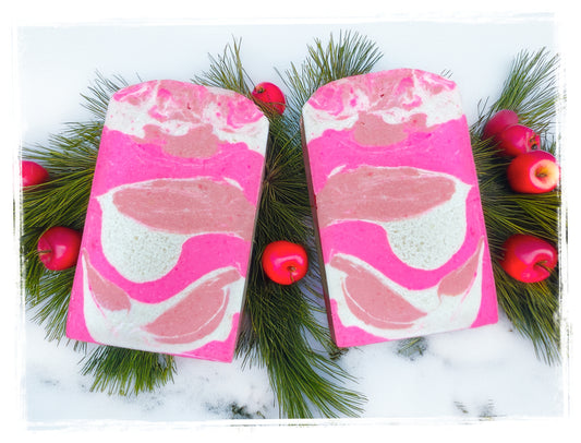 Two Winter Candy Apple goat milk soap bars with bright pink, blush, and white swirls, arranged on pine branches with small red apples.