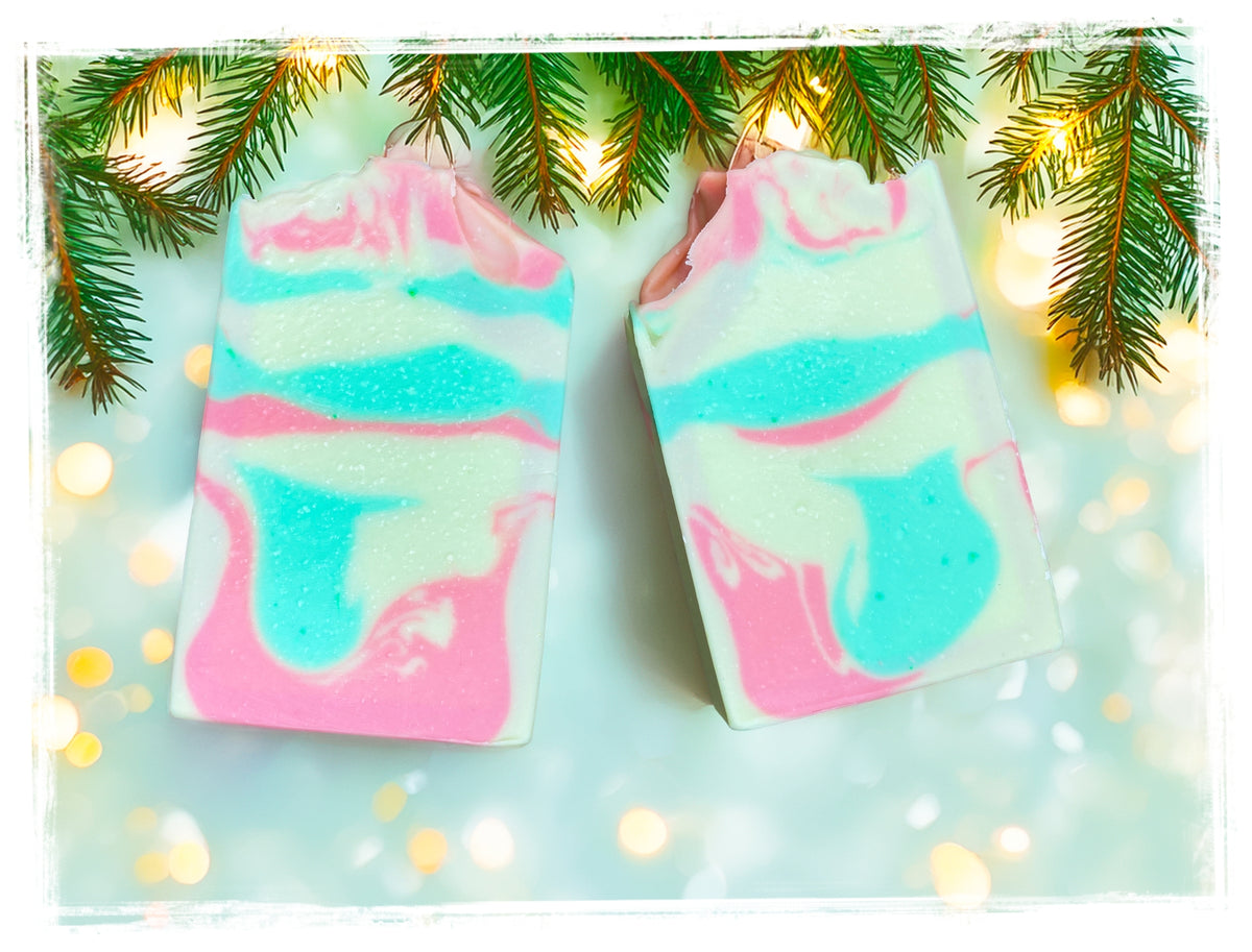 Two Christmas Tree Goat Milk Soap bars with pink, turquoise, and creamy white swirls beneath evergreen branches and warm holiday lights.