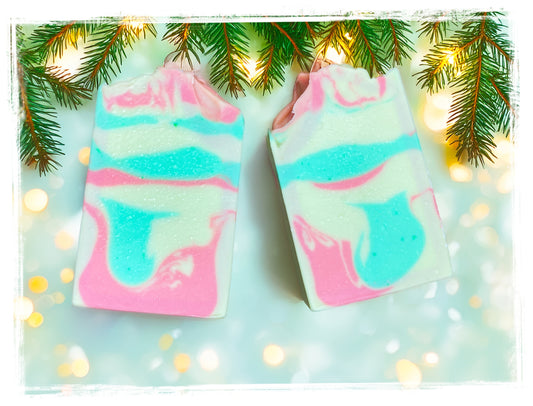 Two Christmas Tree Goat Milk Soap bars with pink, turquoise, and creamy white swirls beneath evergreen branches and warm holiday lights.