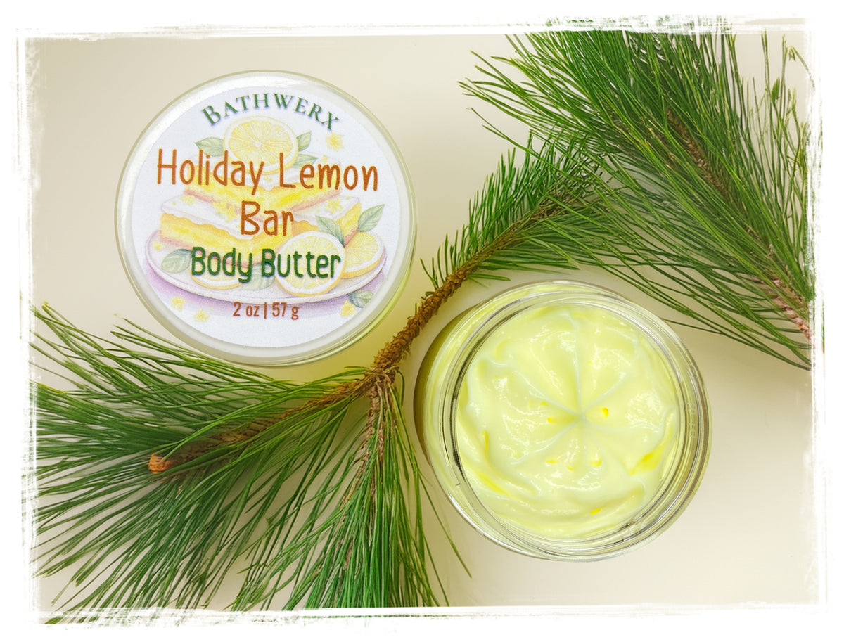 Open jar of pale yellow Holiday Lemon Bar body butter beside its labeled lid and fresh pine branches.