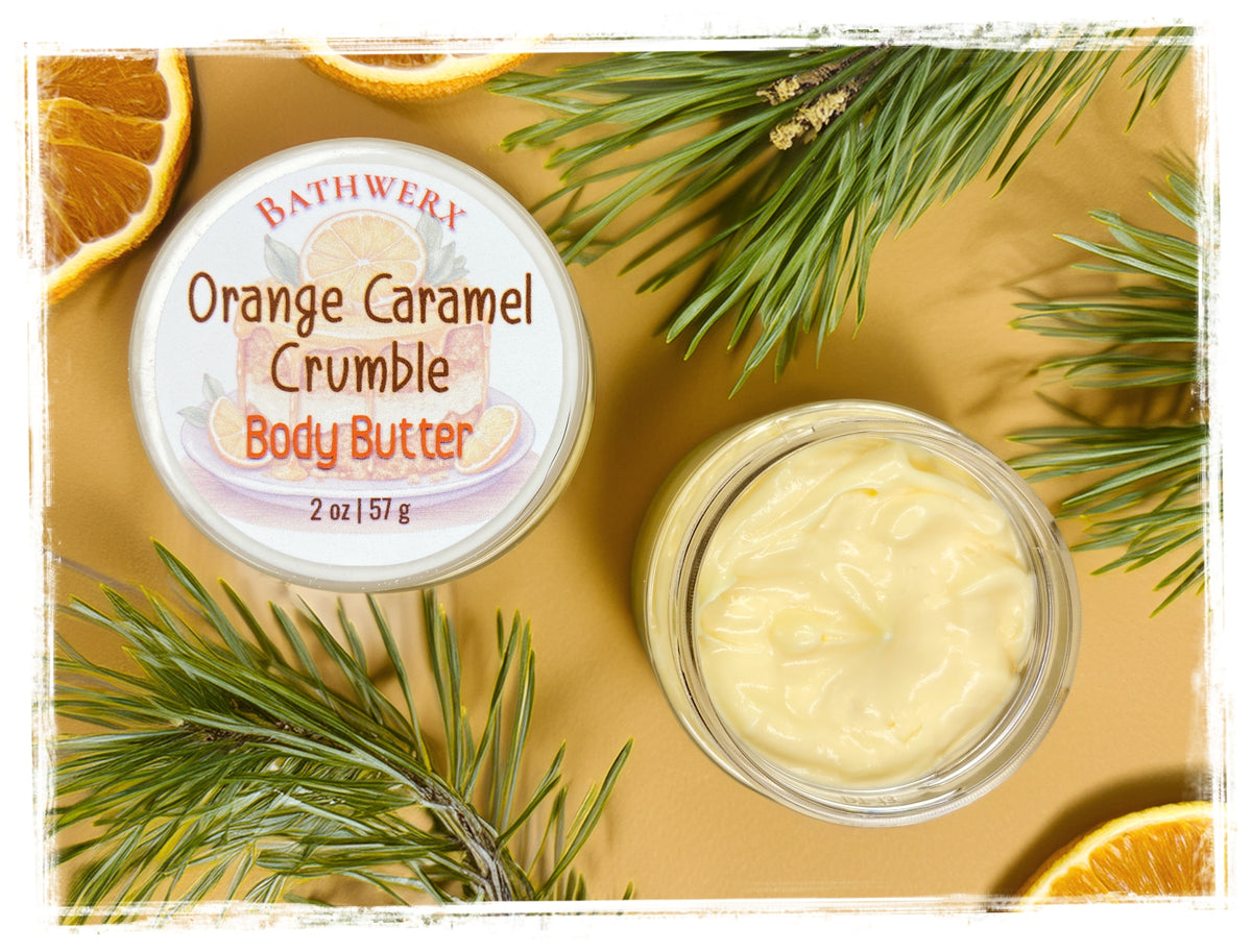 Open jar of Orange Caramel Crumble body butter beside its labeled lid, orange slices, and pine branches.