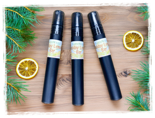 Three black Holiday Lemon Bar perfume spray bottles on a wooden surface with dried lemon slices and evergreen branches.