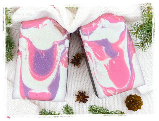 Two Sweater Weather Goat Milk Soap bars with pink, purple, and white swirls on a white knit sweater, surrounded by evergreen sprigs and pinecones.