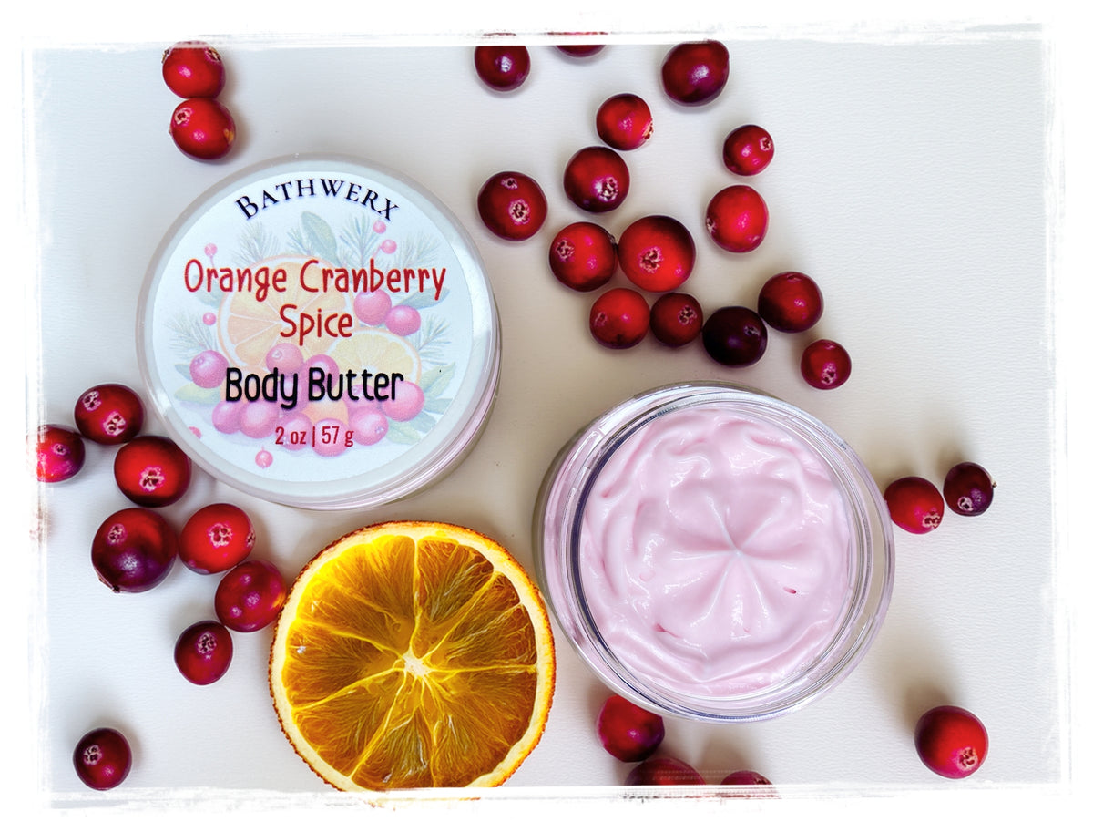Open jar of pink Orange Cranberry Spice body butter beside its labeled lid, cranberries, and an orange slice.