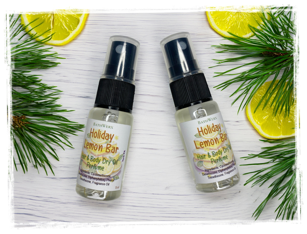 Two Holiday Lemon Bar Hair & Body Dry Oil spray bottles with clear caps, arranged with lemon slices and pine branches on white wood.