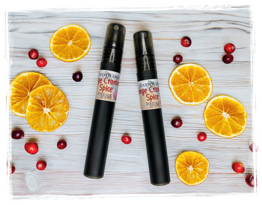 Two black Orange Cranberry Spice perfume spray bottles surrounded by orange slices and cranberries.