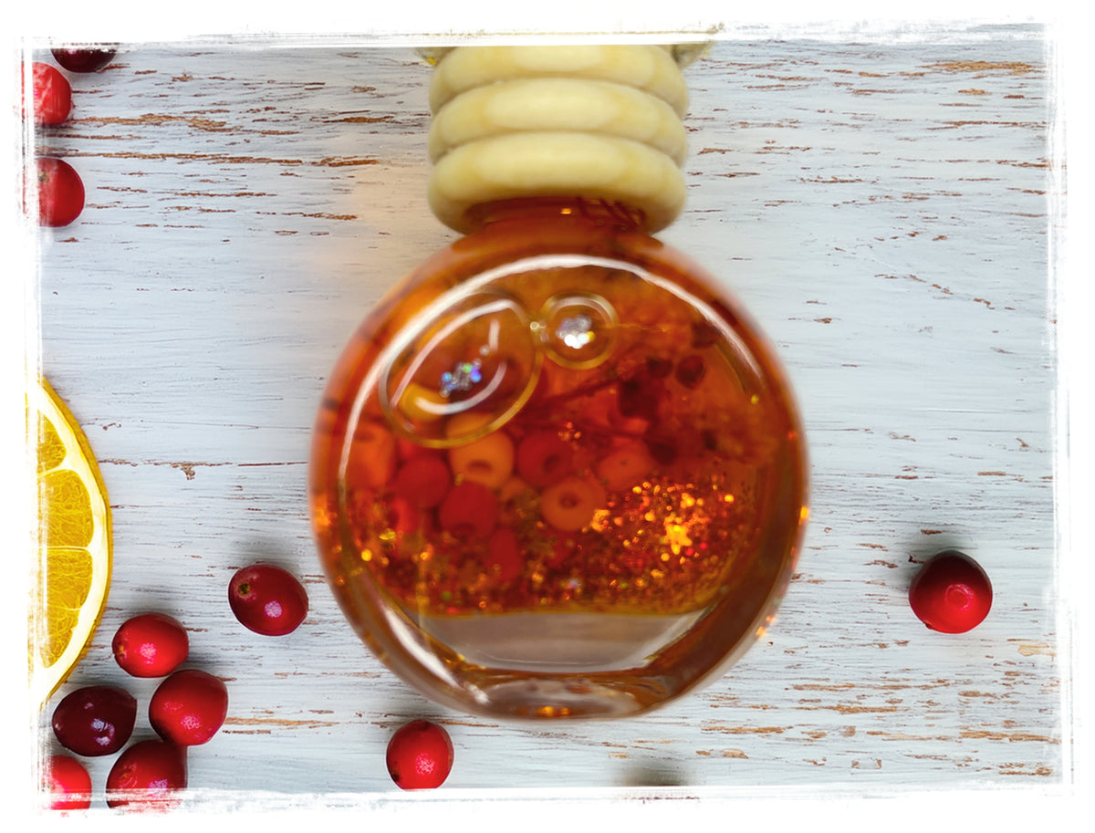 Orange Cranberry Spice car charm with a round glass bottle, wooden cap, orange and red beads, and gold glitter, surrounded by cranberries and an orange slice.