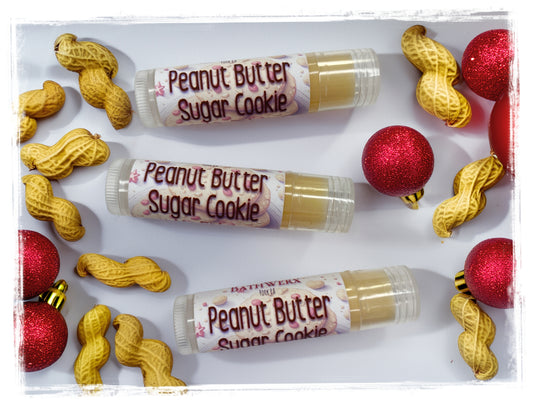 Three tubes of Peanut Butter Sugar Cookie lip balm surrounded by peanuts and red glitter Christmas ornaments.
