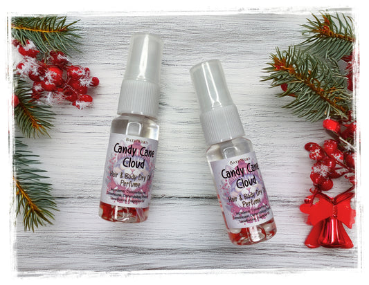 Two Candy Cane Cloud Hair & Body Dry Oil Perfume spray bottles on white wood with evergreen branches and red holiday decorations.