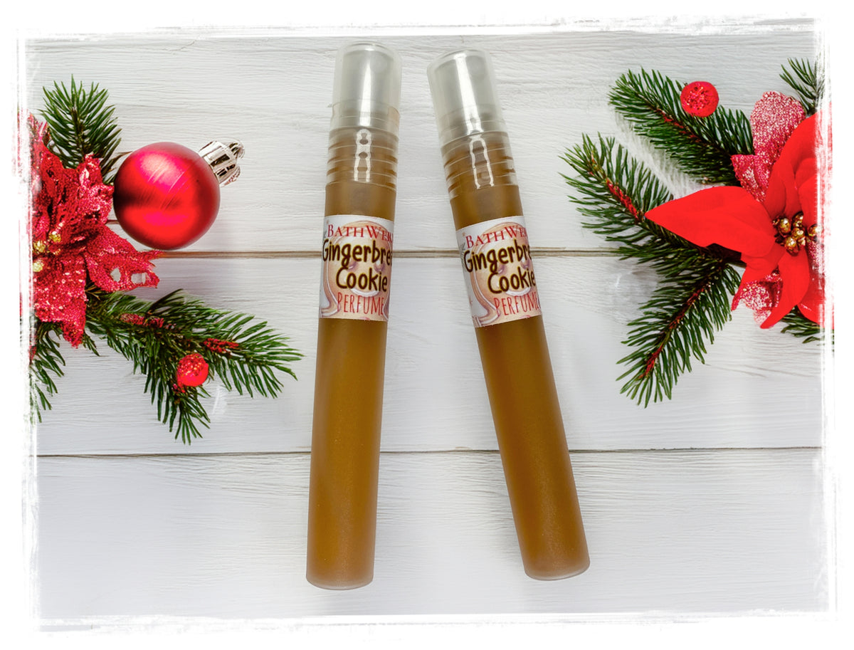 Two Gingerbread Cookie perfume spray bottles with silver caps on white wood, surrounded by evergreen branches and red holiday decorations.