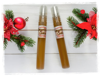 Two Gingerbread Cookie perfume spray bottles with silver caps on white wood, surrounded by evergreen branches and red holiday decorations.