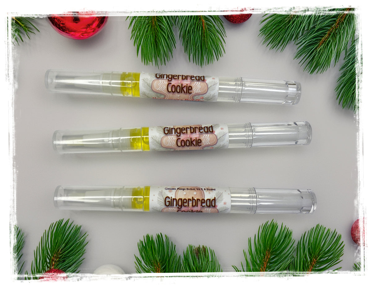 Three Gingerbread Cookie nail and cuticle oil pens with clear caps, surrounded by pine branches and red ornaments.
