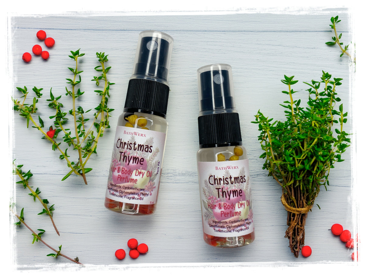 Two Christmas Thyme Hair & Body Dry Oil Perfume spray bottles surrounded by thyme sprigs and red berries on white wood.