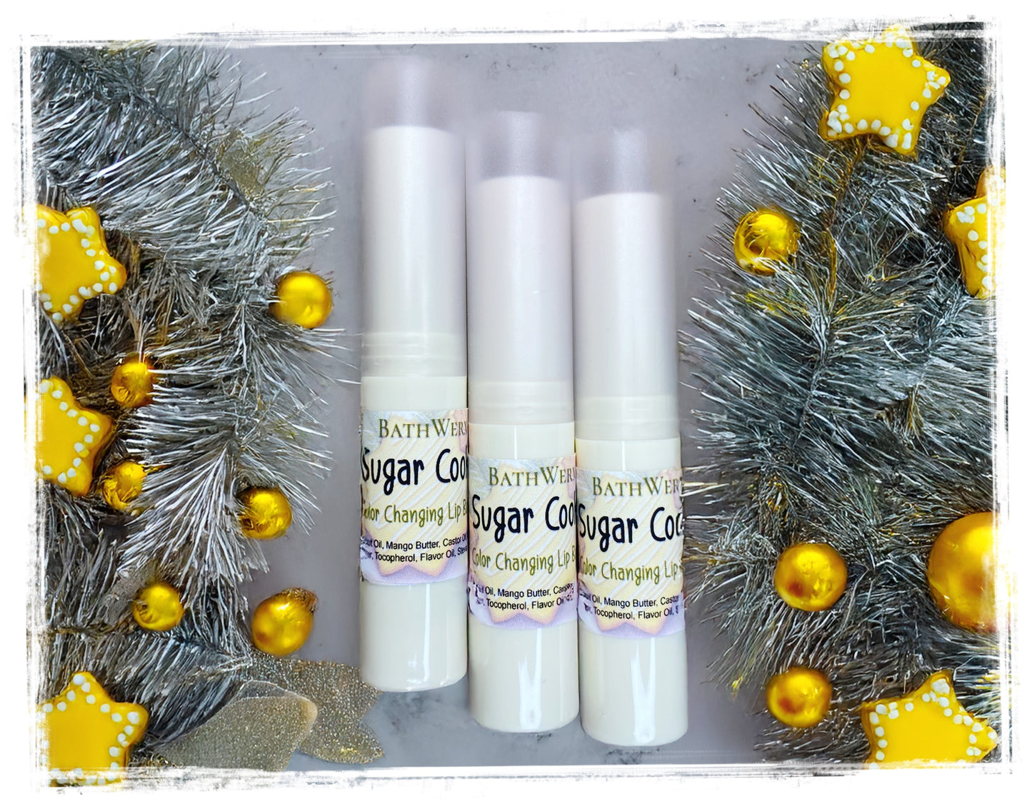 Three Sugar Cookie lip balms with decorative Christmas trees and gold ornaments in the background.