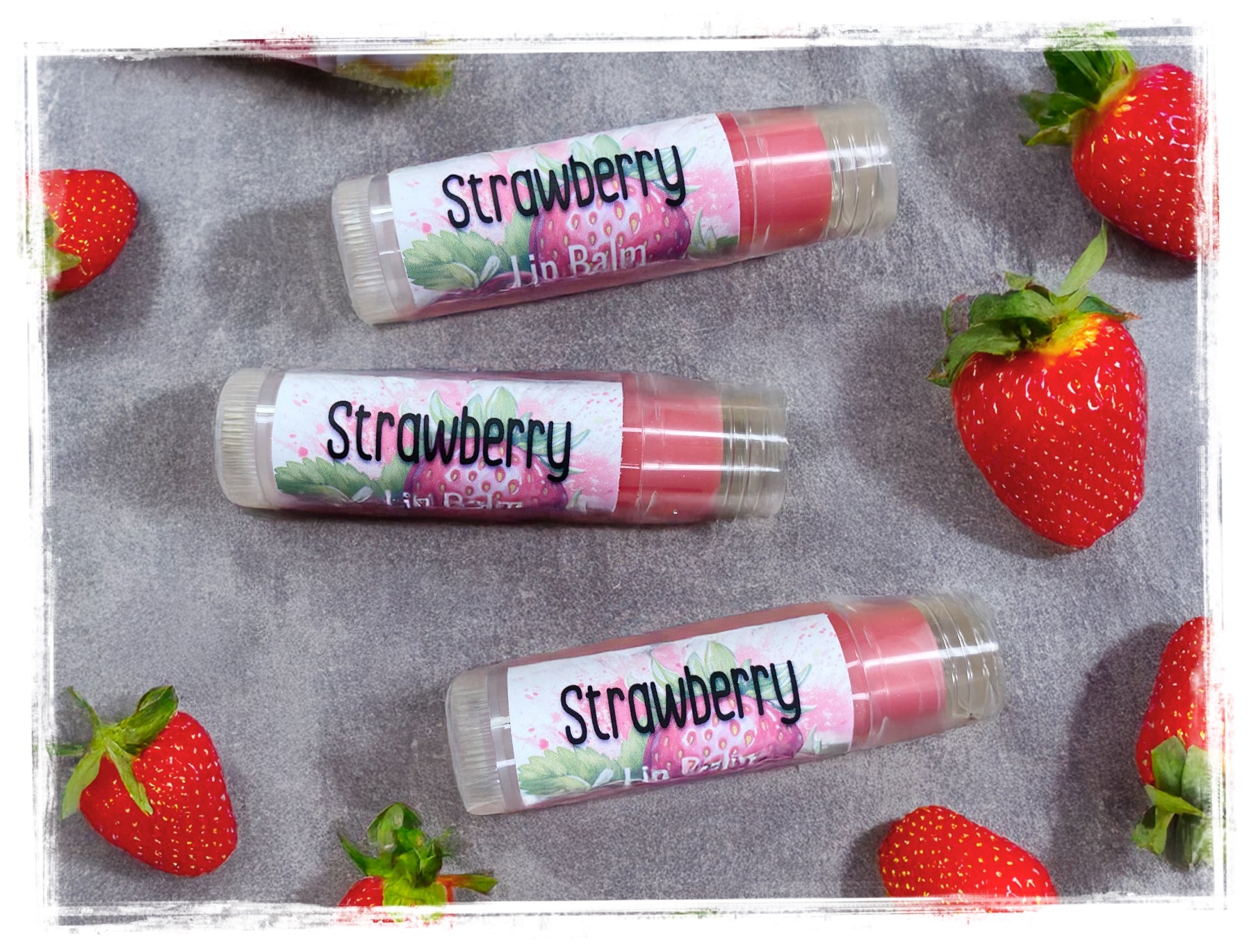 Three strawberry-flavored lip balms on a gray surface with strawberries around them.