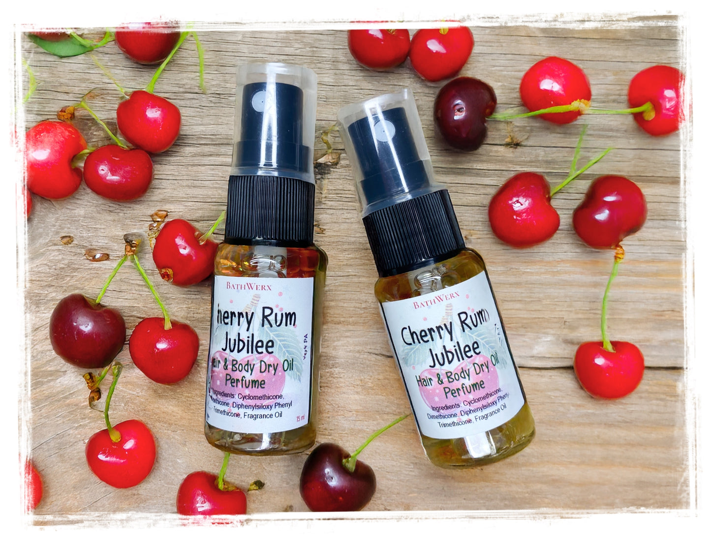 Two bottles of Cherry Rum Jubilee hair and body dry oil perfume surrounded by cherries on a wooden surface.