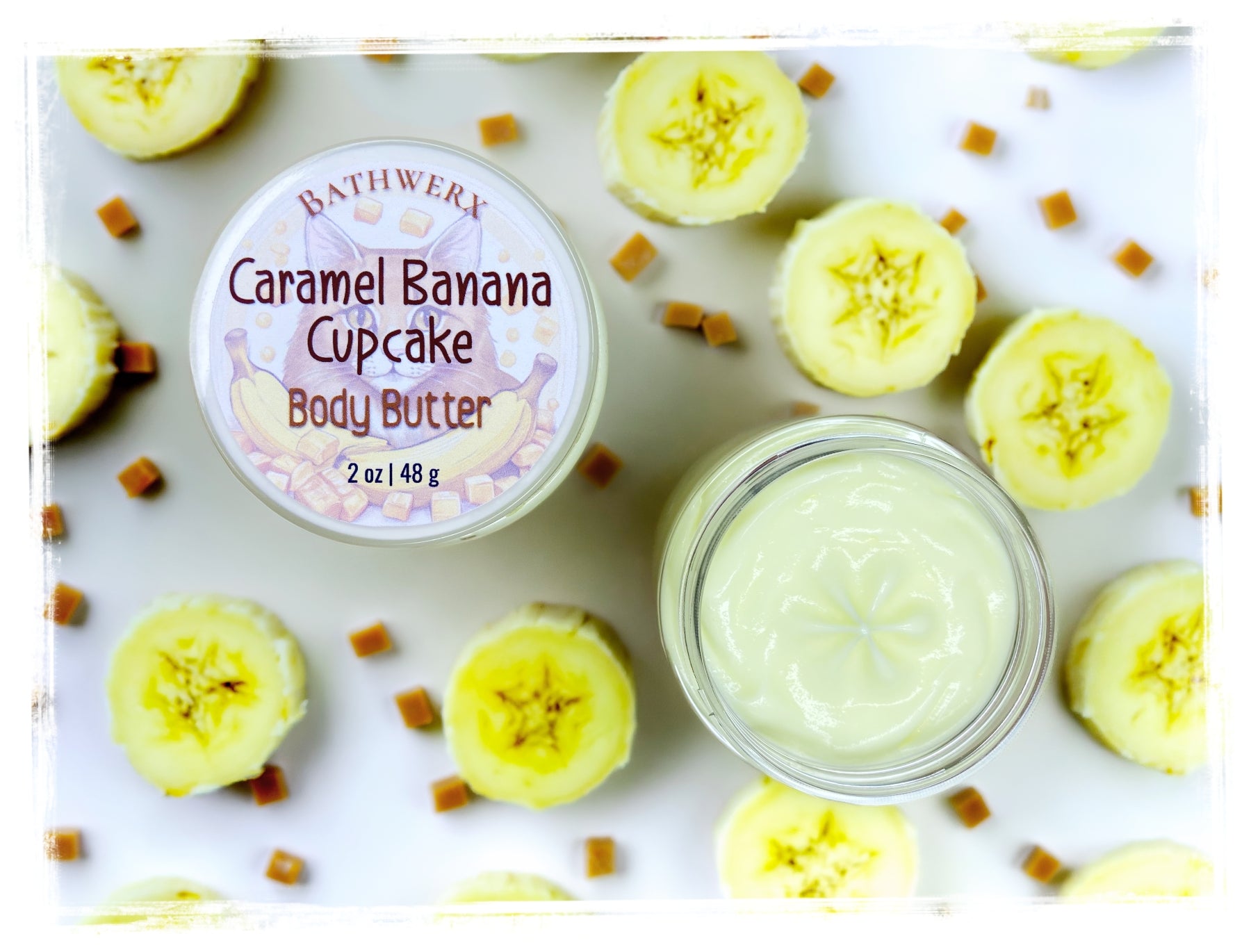 BathWerx Caramel Banana Cupcake Body Butter in a clear glass jar, 2 oz, surrounded by fresh banana slices and soft caramel candy pieces on a white surface