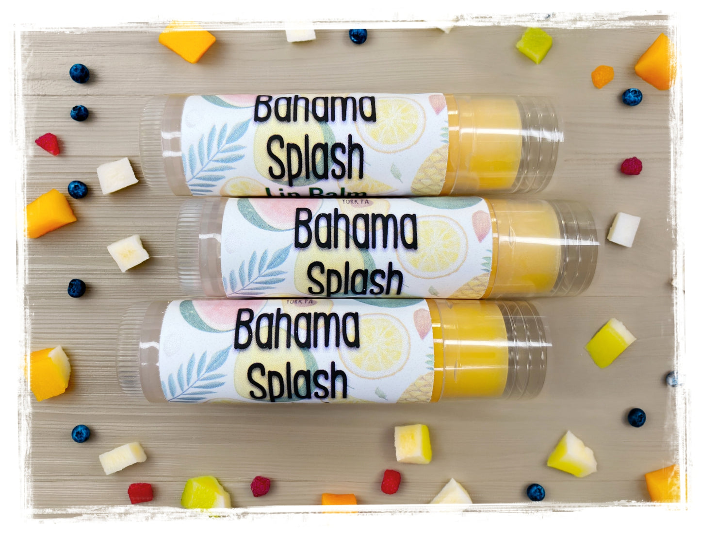 Three BathWerx Bahama Splash Lip Balm tubes in clear packaging with tropical palm leaf labels, surrounded by colorful mixed fruit pieces including mango, blueberries, and coconut on a wood surface