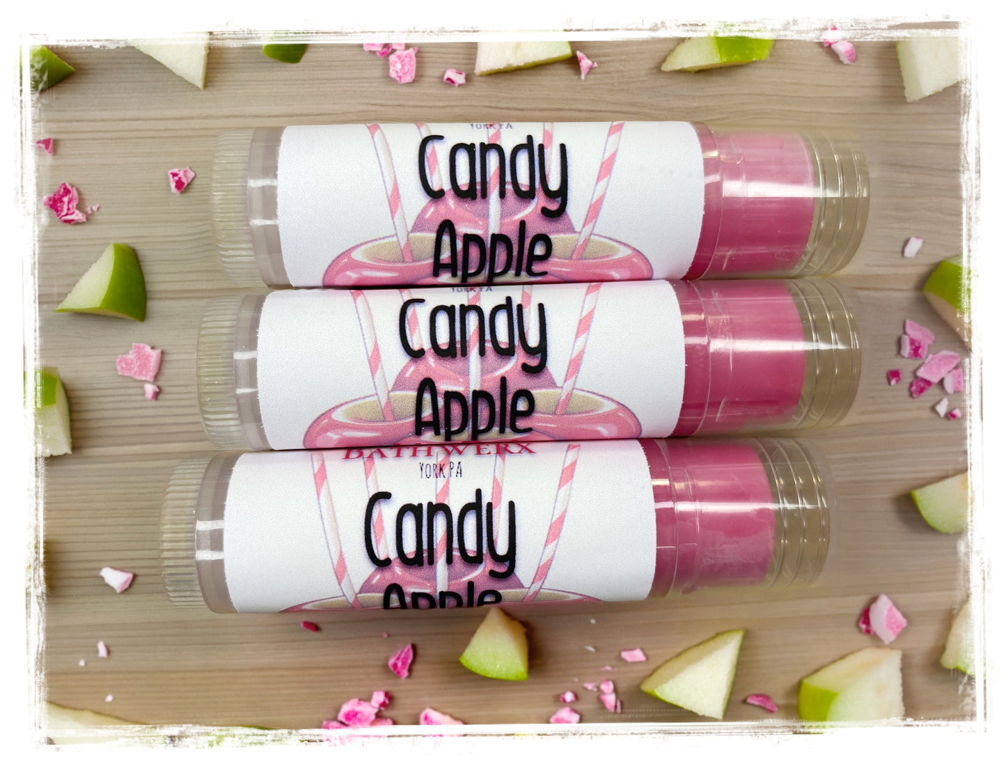 Three BathWerx Candy Apple Lip Balm tubes in clear packaging with pink candy apple labels, surrounded by fresh green apple slices and crushed pink candy on a wood surface
