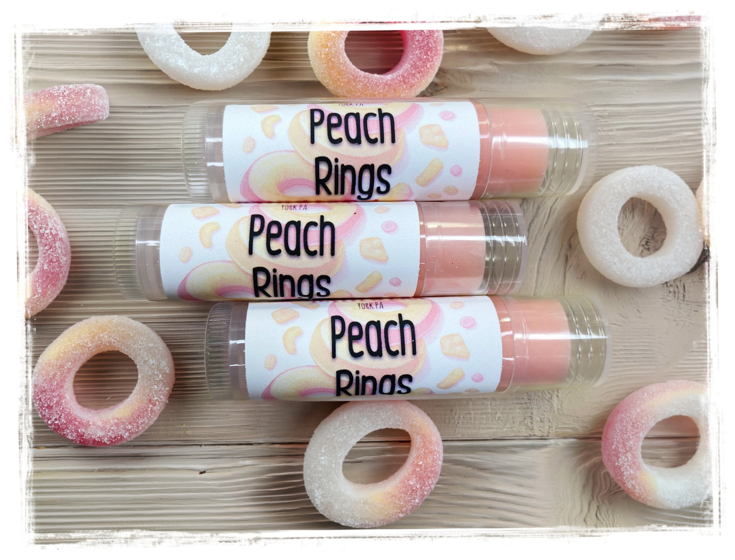 Three BathWerx Peach Rings Lip Balm tubes in clear packaging with peach-illustrated labels, surrounded by sugary pink and white peach ring gummy candies on a wood surface