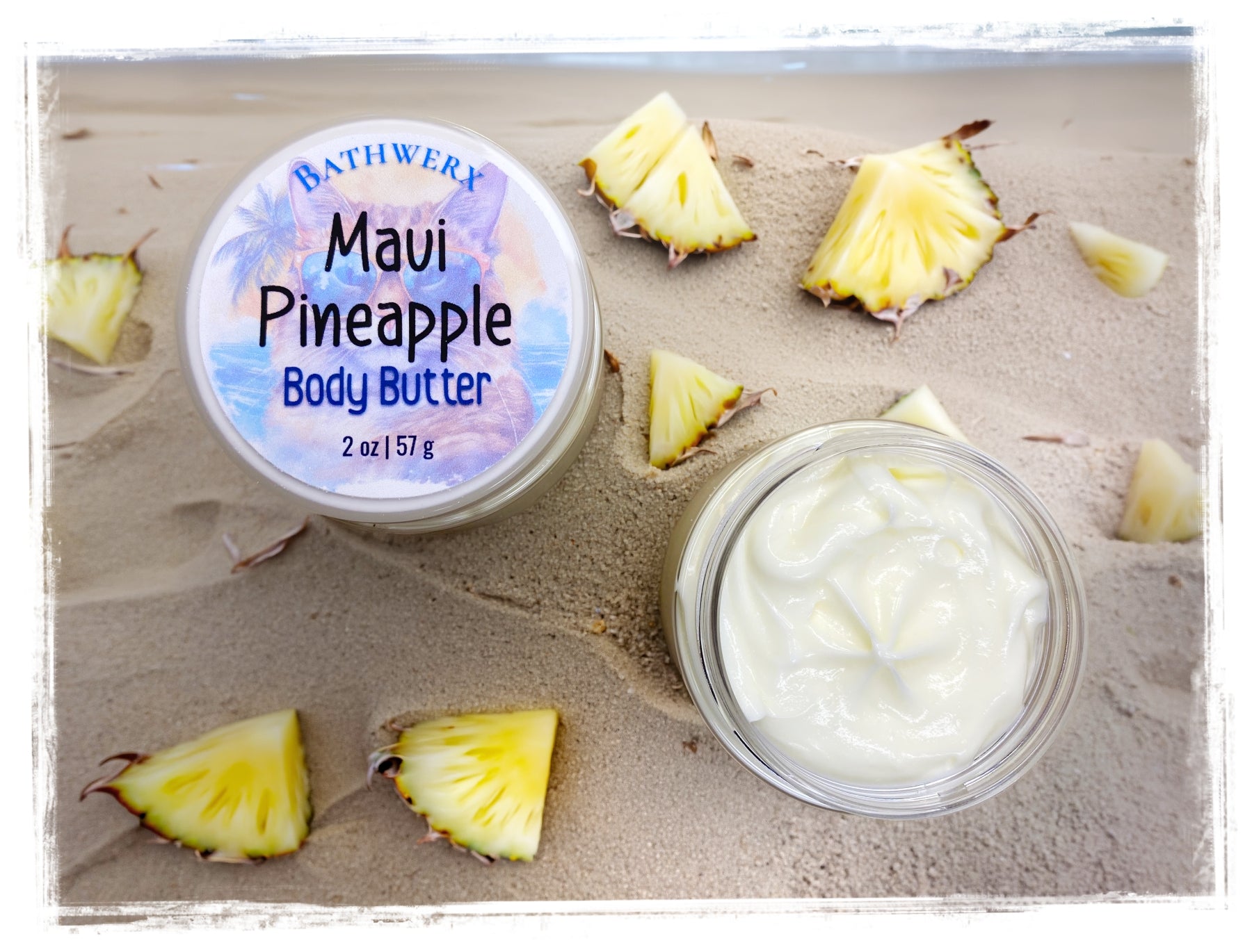 Maui Pineapple Body Butter by BathWerx — open glass jar of whipped body butter and labeled lid displayed on sandy beach surrounded by fresh pineapple slices