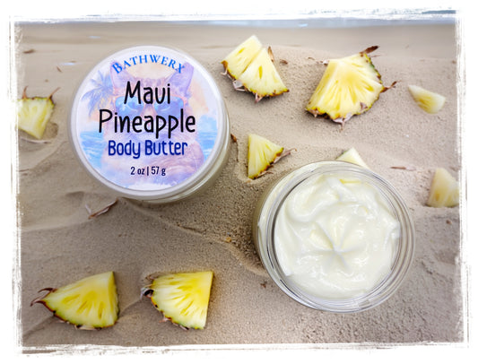 Maui Pineapple Body Butter by BathWerx — open glass jar of whipped body butter and labeled lid displayed on sandy beach surrounded by fresh pineapple slices