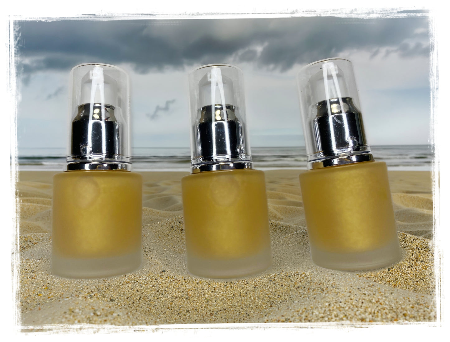 BathWerx Goldburst Body Dry Oil — three frosted gold pump bottles on sandy beach with stormy sky