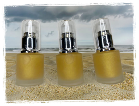 BathWerx Goldburst Body Dry Oil — three frosted gold pump bottles on sandy beach with stormy sky