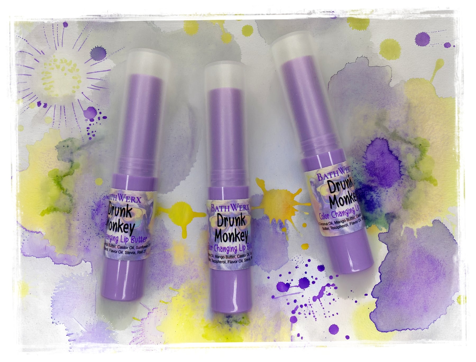 Three BathWerx Drunk Monkey Color-Changing Lip Butter tubes in lavender packaging, arranged on a watercolor-splashed background in purple and yellow